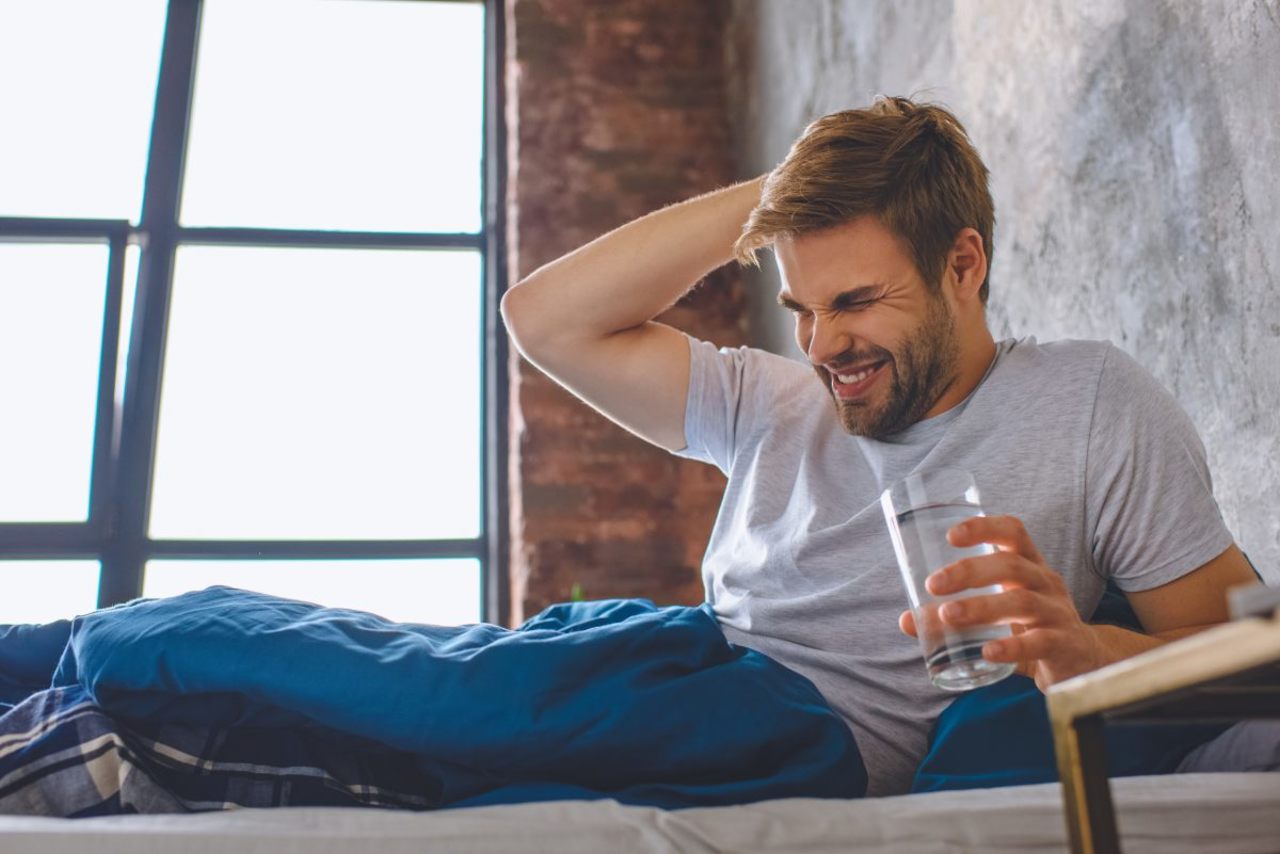 Mann liegt morgens im Bett mit Kopfschmerzen, er hat ein Wasserglas in der Hand.