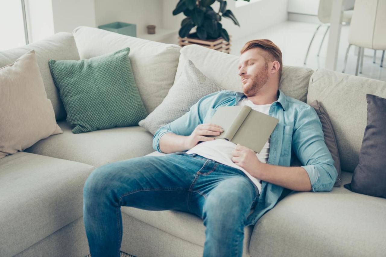 Mann mit Boreout ist gelangweilt und schläft mit Buch auf Sofa.