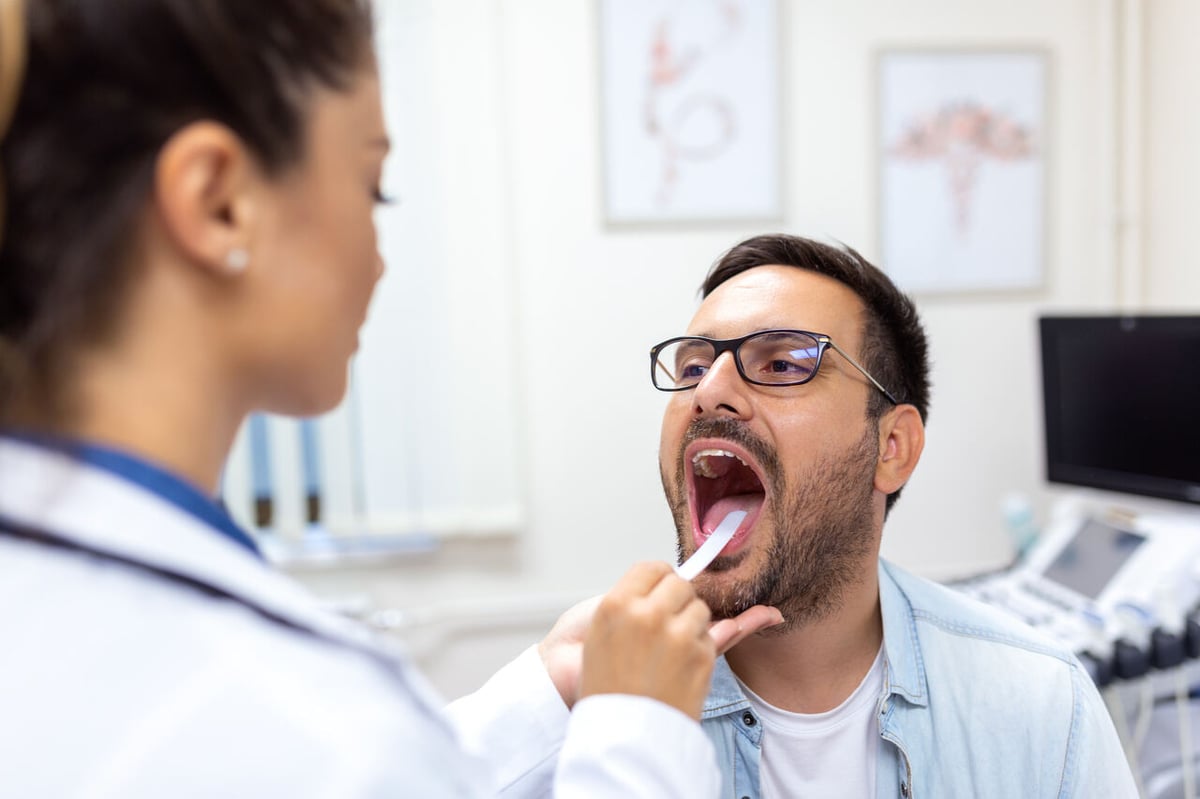 Eine Ärztin drückt bei ihrem Patienten die Zunge mit einem Spatel runter um Hals und Rachen zu untersuchen.