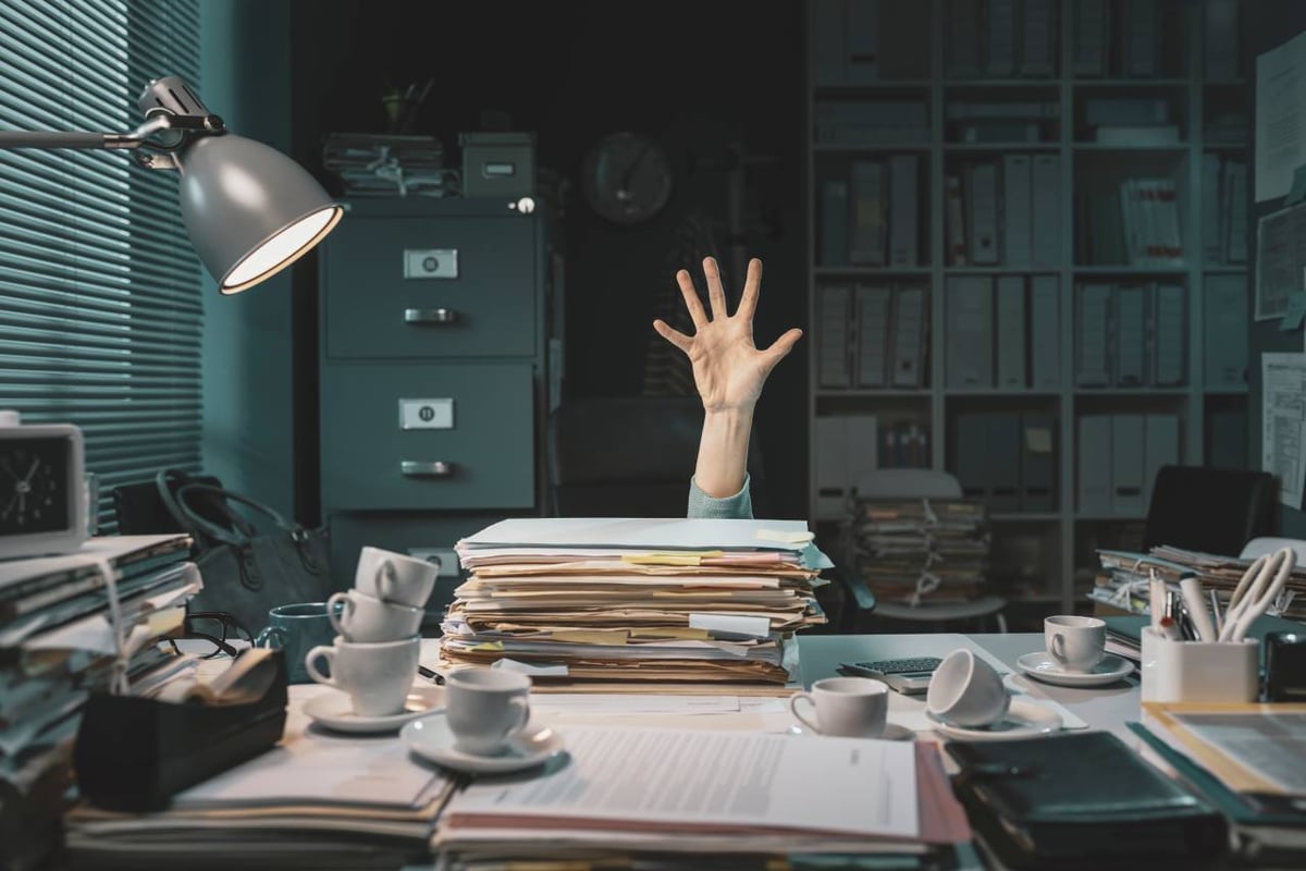 Ein voller Schreibtisch steht in einem Büro. In der Mitte des Bildes ist eine ausgestreckte Hand. Es ist ein Symbolbild für jemanden, der in Arbeit ertrinkt.