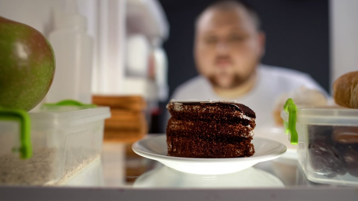 Es ist ein Mann zu sehen, der die Kühlschranktür öffnet. Im Fokus des Bildes ist ein Schokokuchen.