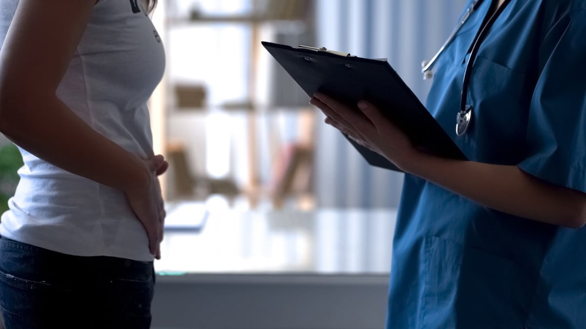 Frau hält sich den Bauch. Ihr gegenüber steht ein Arzt mit einem Klemmbrett.