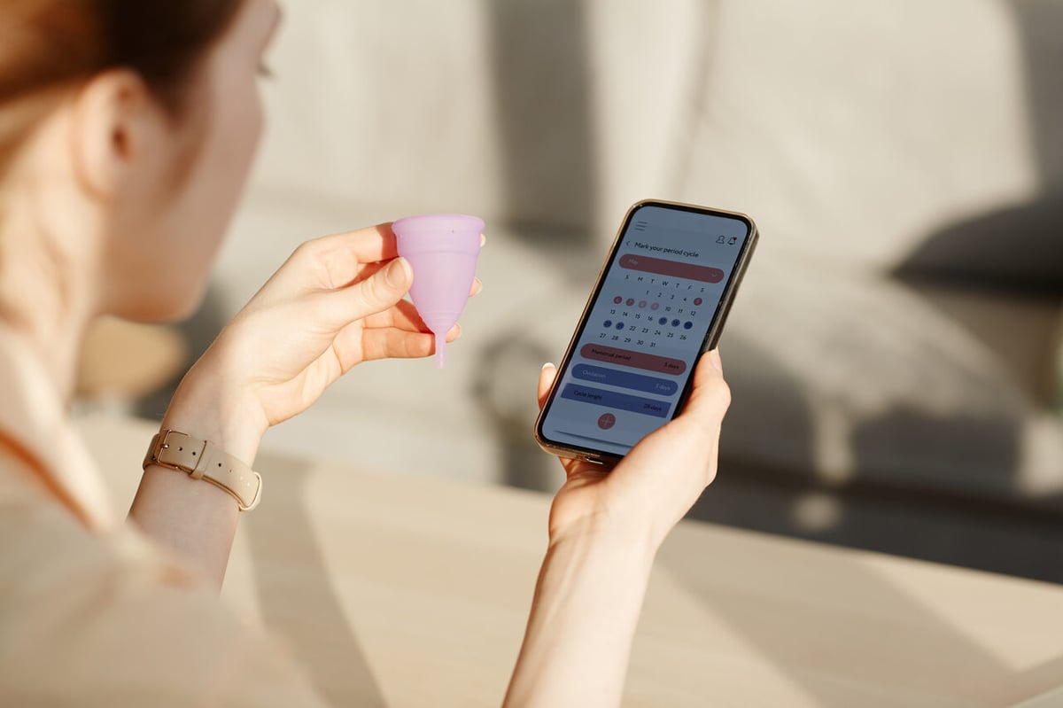 Frau hält Menstruationstasse in der linken Hand und Handy mit Zykluskalender in der rechten.