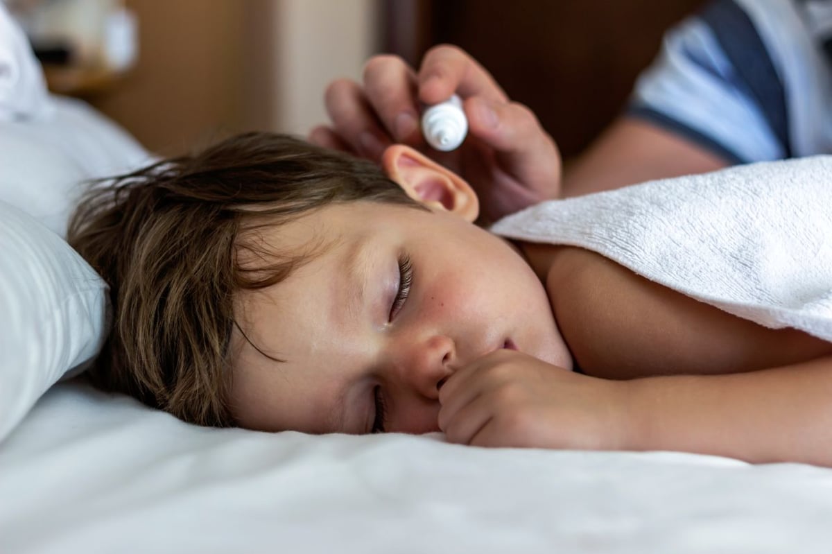 Kind mit Ohrenschmerzen liegt im Bett und bekommt Ohrentropfen verabreicht.