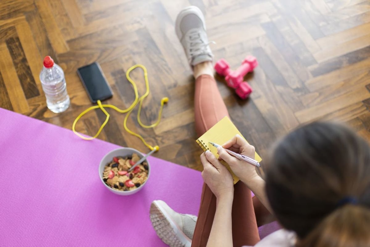 Kombination aus Ernährung und Sport um abzunehmen. Junge Frau schreibt in ein Notizbuch, sitzt auf Yogamatte und hat gesundes Müsli und Hanteln vor sich.