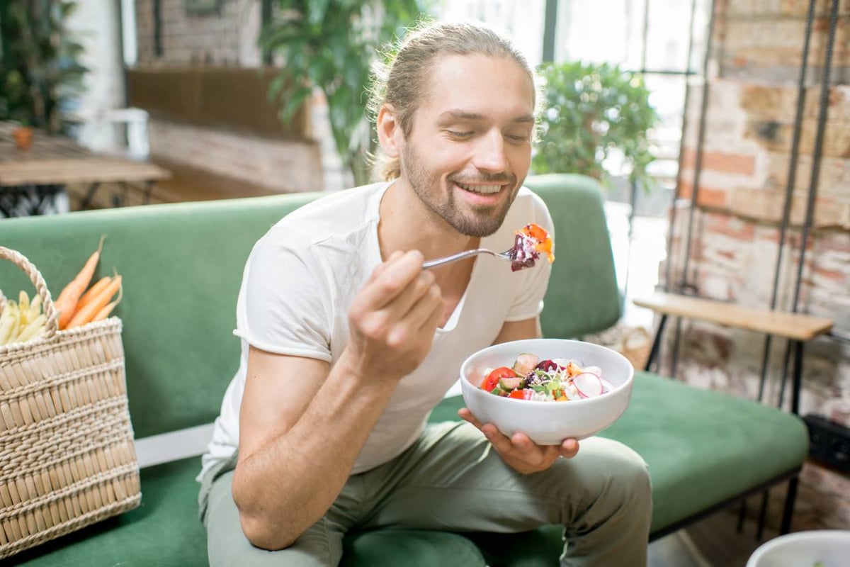 Auf dem Bild ist ein junger Mann zu sehen, der auf einem grünem Sofa sitzt und einen Salat isst.