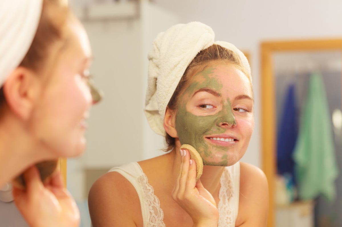 Junge Frau vor dem Spiegel im Badezimmer mit einer Gesichtsmaske aus Heilerde.