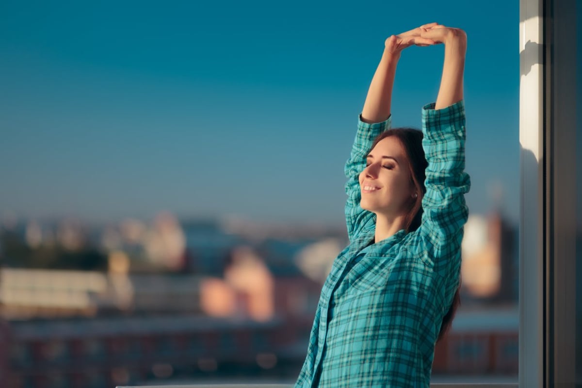 Junge Frau mit kariertem Hemd streckt sich in der Sonne. Im Hintergrund ist die Skyline einer Stadt zu sehen.