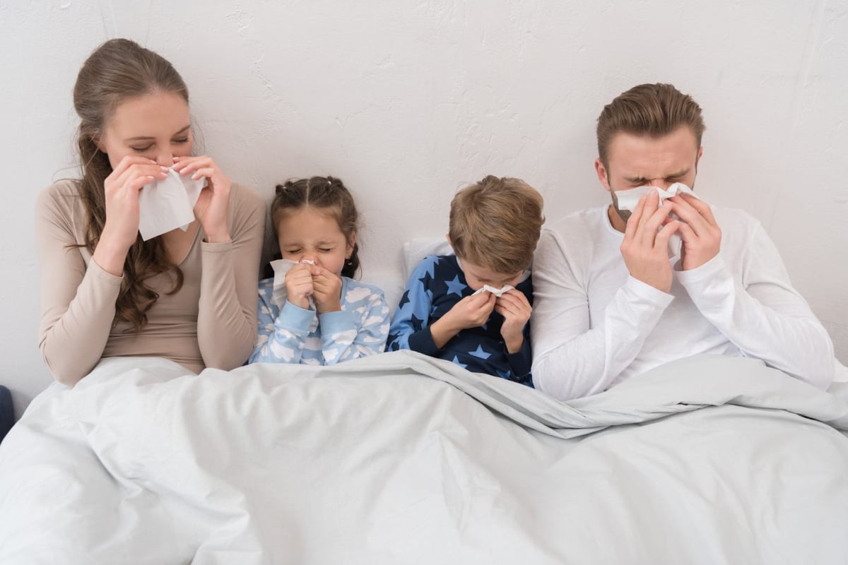 Familie ist zusammen im Bett. Alle sind krank und schnäuzen in ein Taschentuch.