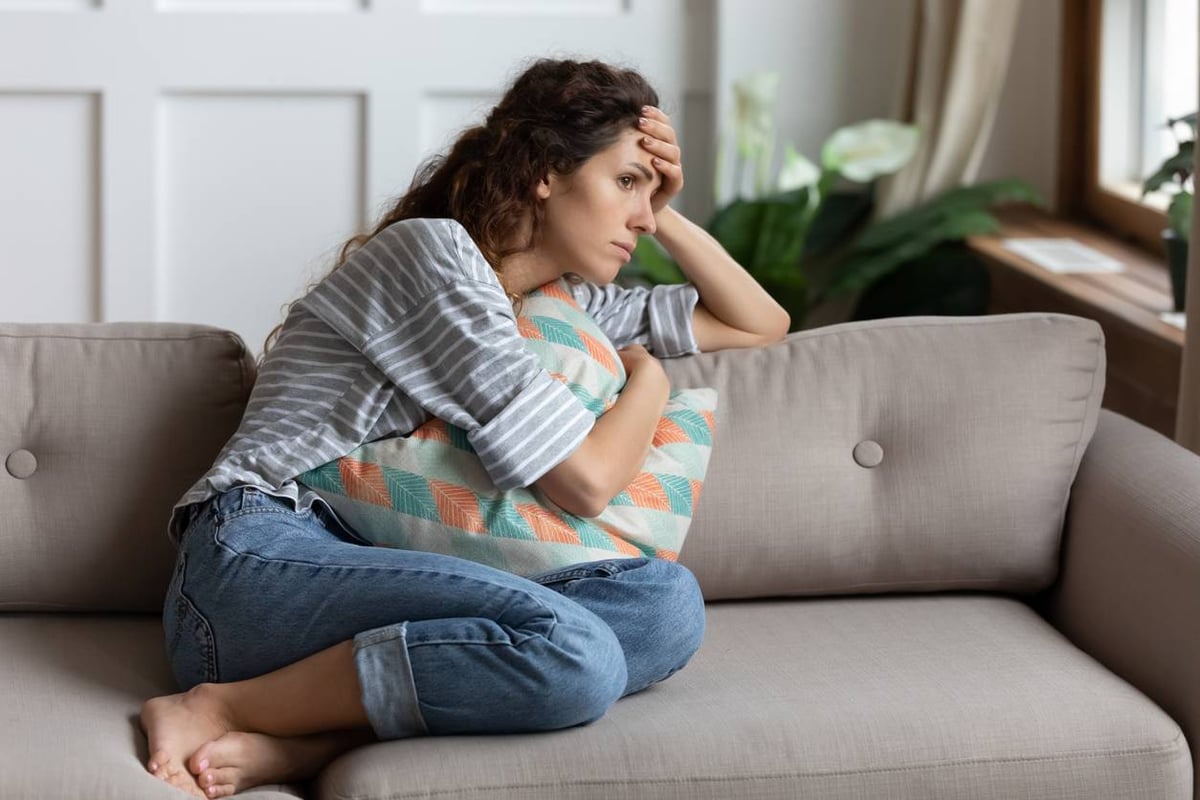 Frau sitzt auf dem Sofa und hält sich die Stirn mit einer Hand und ein Kissen mit der anderen Hand