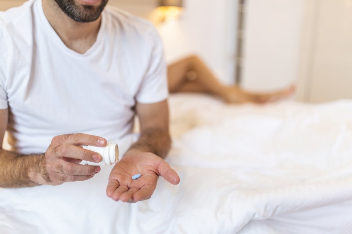 Mann sitzt auf Bett mit Viagra Pille auf der Hand.