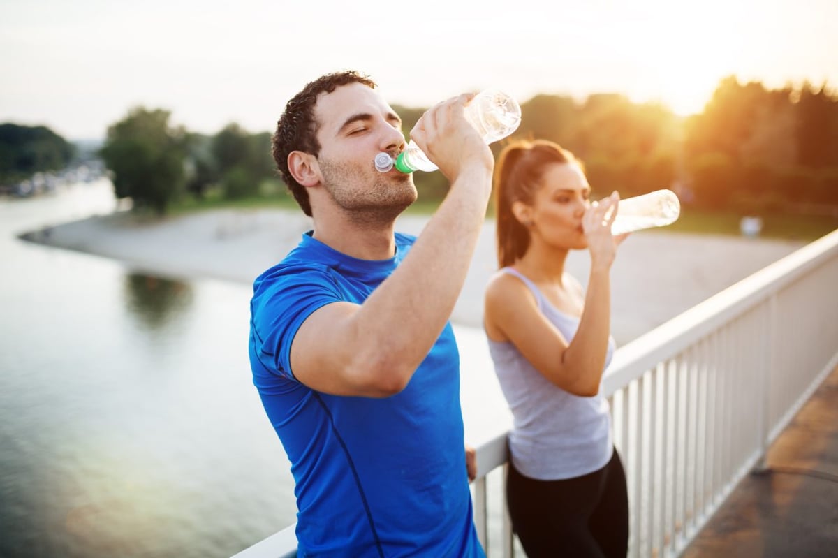 Es ist ein junges Paar in Sport-Outfits zu sehen, beide trinken Wasser.