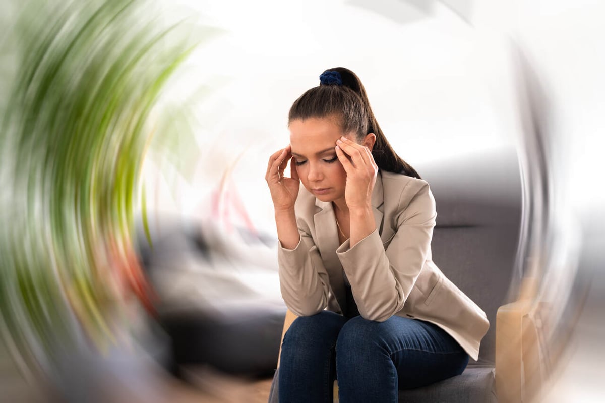 Eine Frau sitzt auf einem Sofa und hält ihren Kopf in den Händen. Ein kreisförmig verschwommener Hintergrund symbolisiert Schwindel.