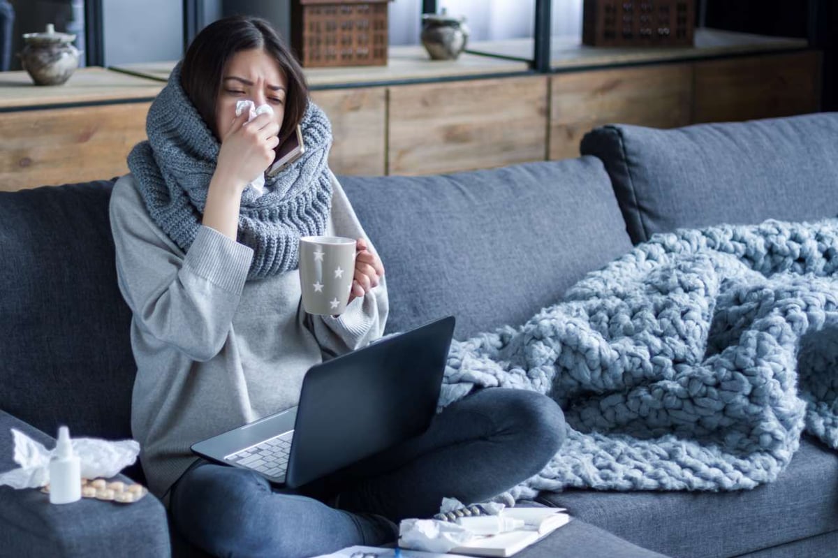 Junge Frau sitzt auf der Couch und putzt sich die Nase. Auf ihrem Schoß steht ein Laptop.