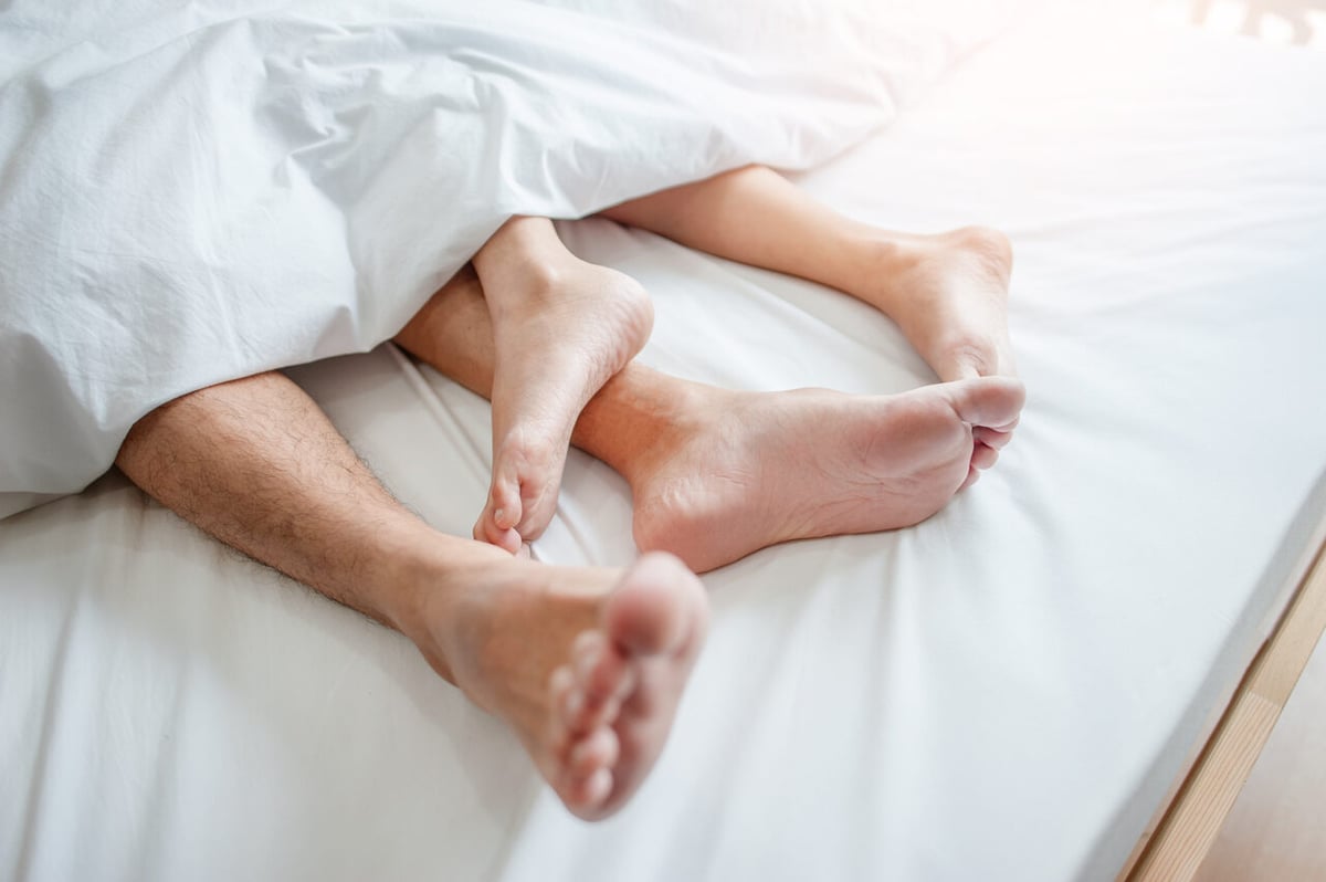 In einem weiß bezogenen Bett gucken die Füße von einem Mann und einer Frau unten aus der Bettdecke.