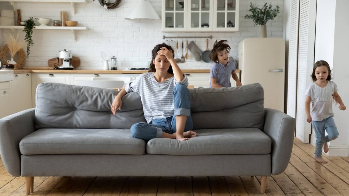 Eine Frau sitzt genervt auf dem Sofa, während Kinder um sie herumtoben.