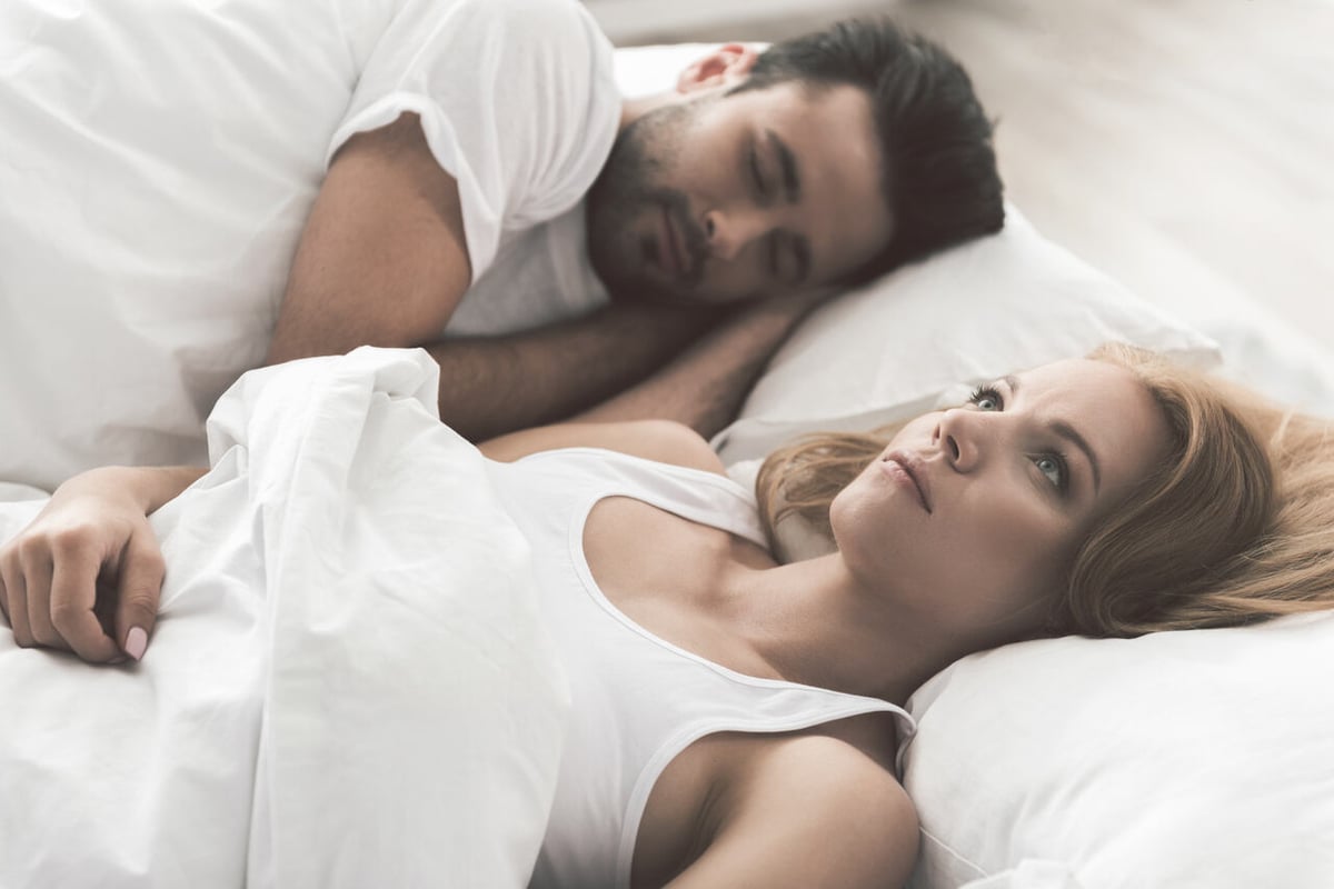 EIne junge Frau liegt wach im Bett und starrt nach oben, daneben schläft ein Mann.
