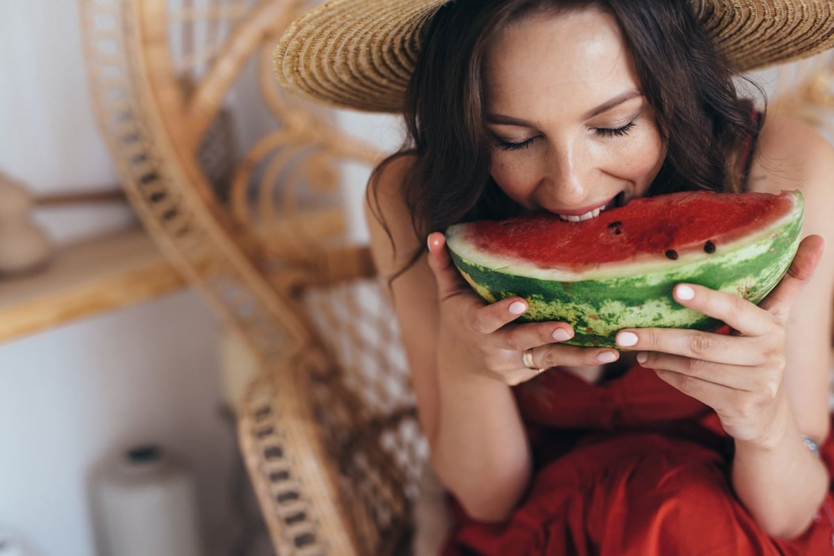 Es ist eine junge Frau mit dunklen Haaren zu sehen, die einen Strohhut trägt und in ein großes Stück Melone beist.