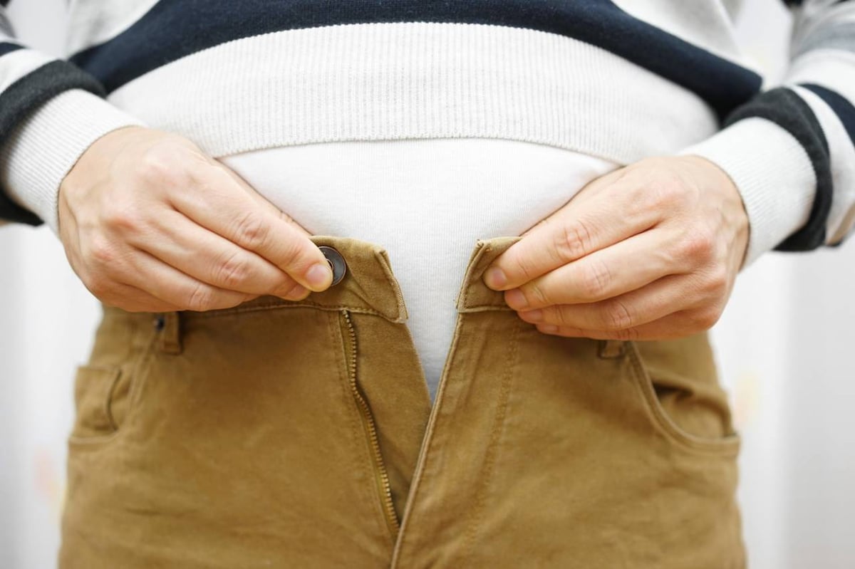 Person bekommt Knopf an der braunen Hose über dem Bauch nicht geschlossen.