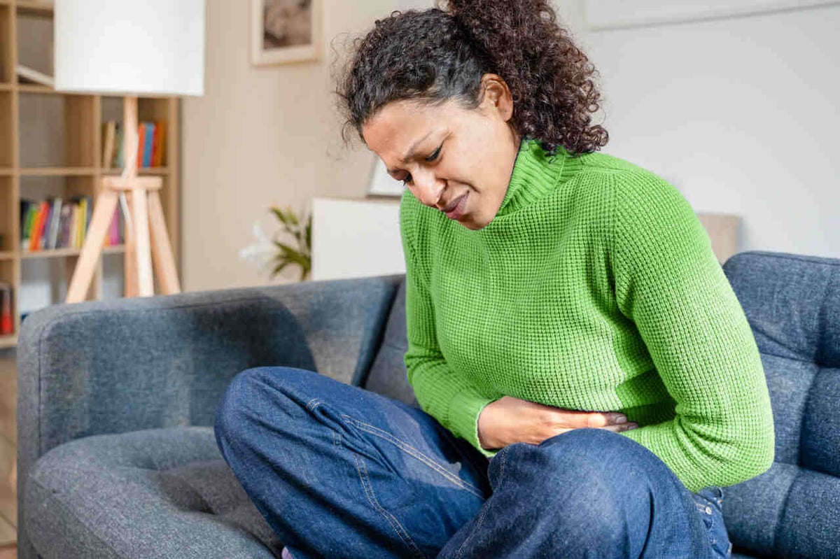 Frau sitzt zusammengekrümmt auf dem Sofa und hält sich den schmerzenden Bauch.