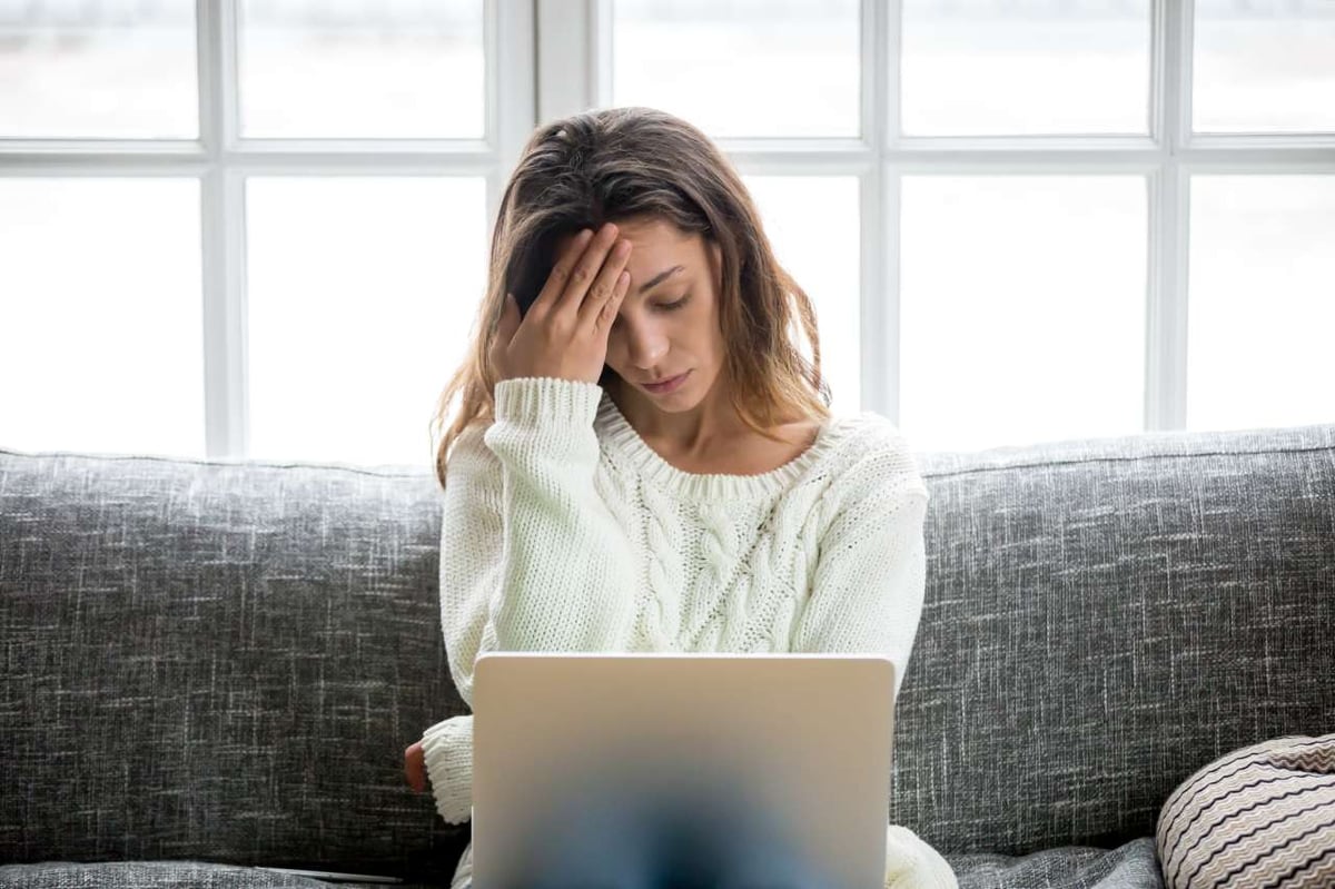 Eine Frau sitzt mit Laptop auf dem Sofa und hat Kopfschmerzen.