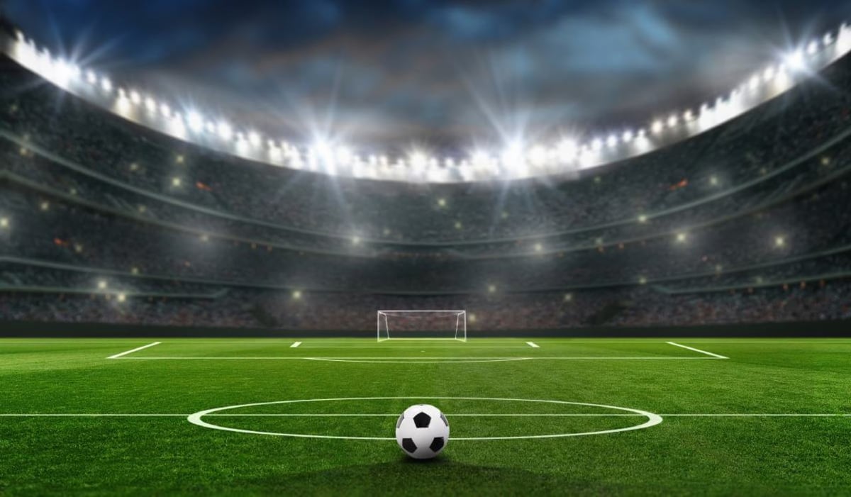 Fussball liegt im Stadion vor der Mittellinie. Im Hintergrund Flutlicht und ein leeres Tor.