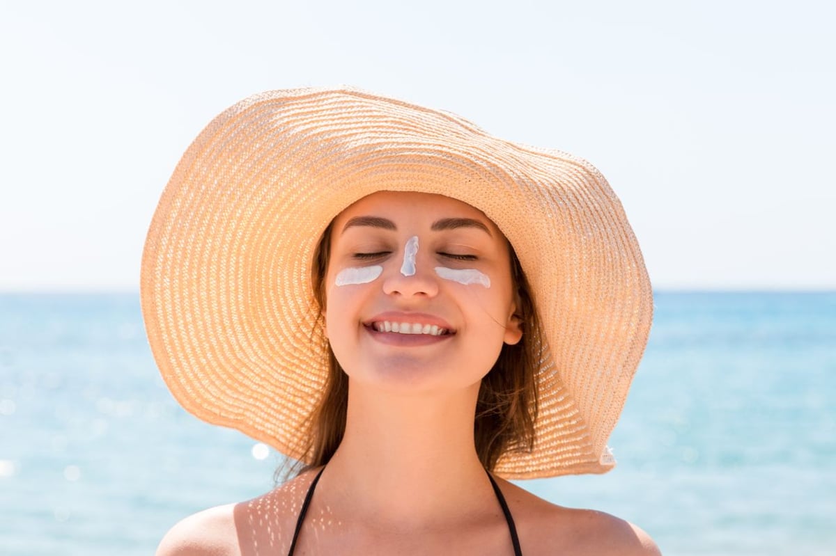 Junge Frau am Meer, die einen Sonnenhut trägt und Sonnencreme im Gesicht hat.