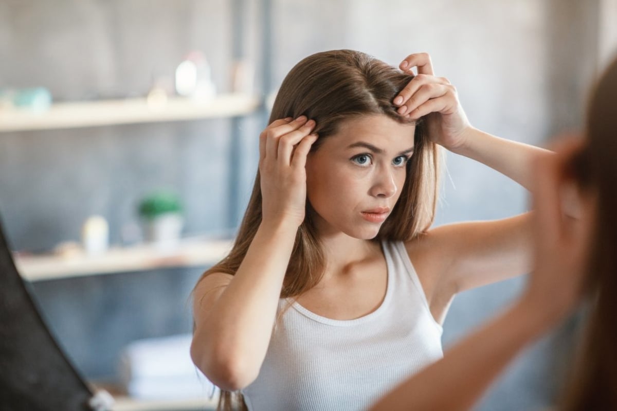 Junge Frau betrachtet sich im Spiegel, sie prüft ihren Haaransatz auf Haarausfall.