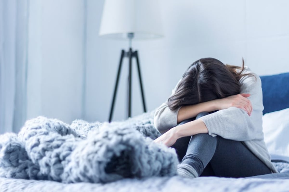 Junge Frau in grauer Kleidung sitzt zusammengerollt auf dem Bett zu Hause. 