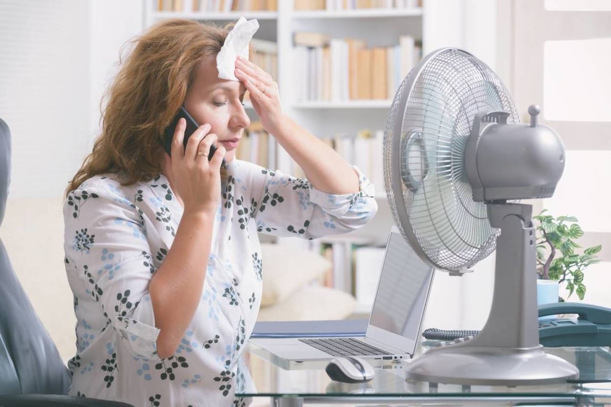 Frau am Telefon sitzt vor dem Ventilator und tupft sich die Stirn mit einem Tuch.