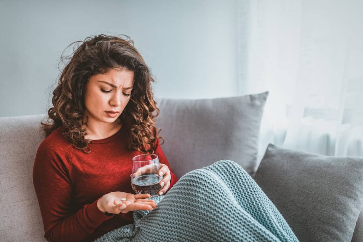Eine Frau sitzt auf dem Sofa und sieht beunruhigt aus. Sie hält ein Glas Wasser und eine Tablette in den Händen.