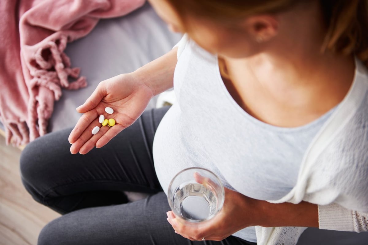 Eine schwangere Frau hält Nahrungsergänzungsmittel in einer Hand und ein Glas Wasser in der anderen.