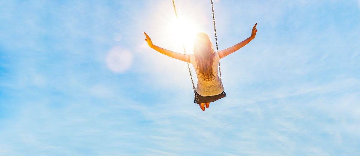 Frau schaukelt vor blauem Himmel und Sonne, sie streckt die Arme zur Seite.