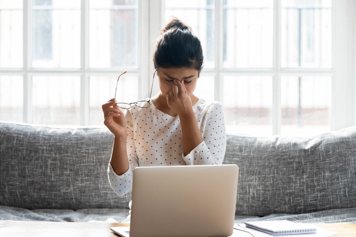 Eine junge Frau sitzt auf der Couch, vor ihr steht ein Laptop. Sie hat ihre Brille abgenommen, schließt die Augen und wirkt gestresst.