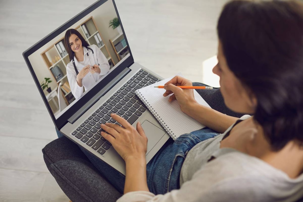 Eine Frau sitzt am Laptop und macht sich während der Online-Sprechstunde mit einer Ärztin Notizen.