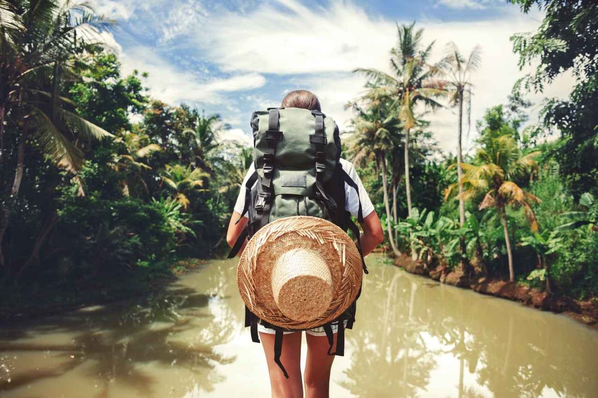 Frau macht einen Abenteuerurlaub. Sie steht mit Rucksack und Strohhut an einem Fluss.