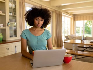 Junge Frau in blauen T-Shirt sitzt in der Küche vor ihrem Laptop.