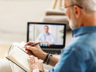 Älterer Mann macht sich Notizen in einem Buch. Im Hintergrund steht ein Laptop, auf dem eine Videosprechstunde läuft.