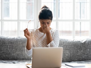 Eine junge Frau sitzt auf der Couch, vor ihr steht ein Laptop. Sie hat ihre Brille abgenommen, schließt die Augen und wirkt gestresst.