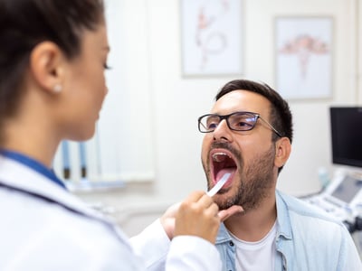 Eine Ärztin drückt bei ihrem Patienten die Zunge mit einem Spatel runter um Hals und Rachen zu untersuchen.