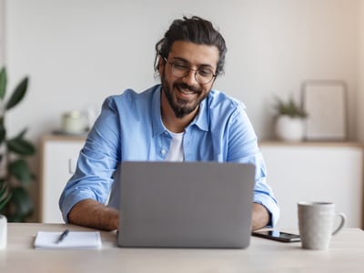 Junger Mann mit dunklen, längeren Haaren und hellblauen Hemd sitzt vor Laptop.