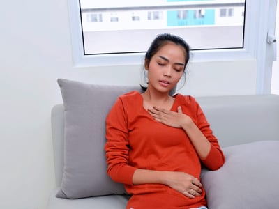 Junge Frau sitzt mit Brustschmerzen und Atemnot auf dem Sofa.