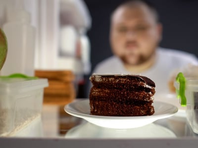 Es ist ein Mann zu sehen, der die Kühlschranktür öffnet. Im Fokus des Bildes ist ein Schokokuchen.