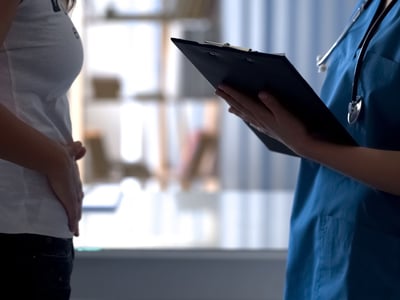 Frau hält sich den Bauch. Ihr gegenüber steht ein Arzt mit einem Klemmbrett.