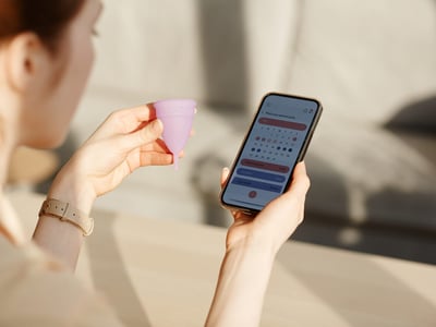 Frau hält Menstruationstasse in der linken Hand und Handy mit Zykluskalender in der rechten.