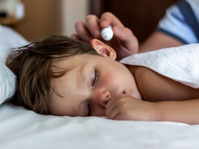 Kind mit Ohrenschmerzen liegt im Bett und bekommt Ohrentropfen verabreicht.