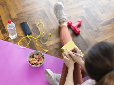 Kombination aus Ernährung und Sport um abzunehmen. Junge Frau schreibt in ein Notizbuch, sitzt auf Yogamatte und hat gesundes Müsli und Hanteln vor sich.