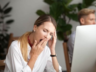 Eine Frau ist müde im Büro und gähnt an ihrem Schreibtisch am Laptop.