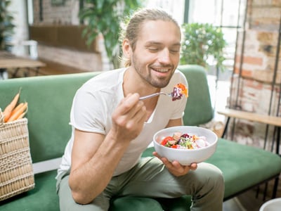 Auf dem Bild ist ein junger Mann zu sehen, der auf einem grünem Sofa sitzt und einen Salat isst.
