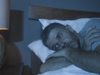 Ein Mann liegt in einem abgedunkelten Raum schlaflos im Bett.